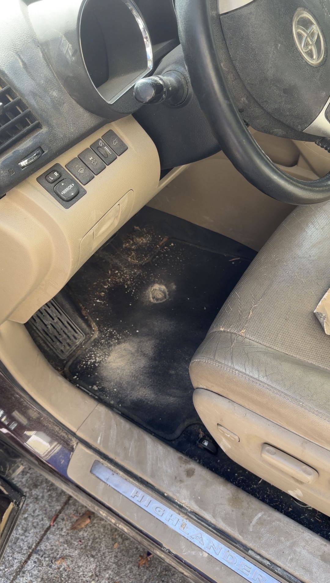 Driver footwell of a Toyota Highlander with pale dust on the mat, sill plate and tan leather seat