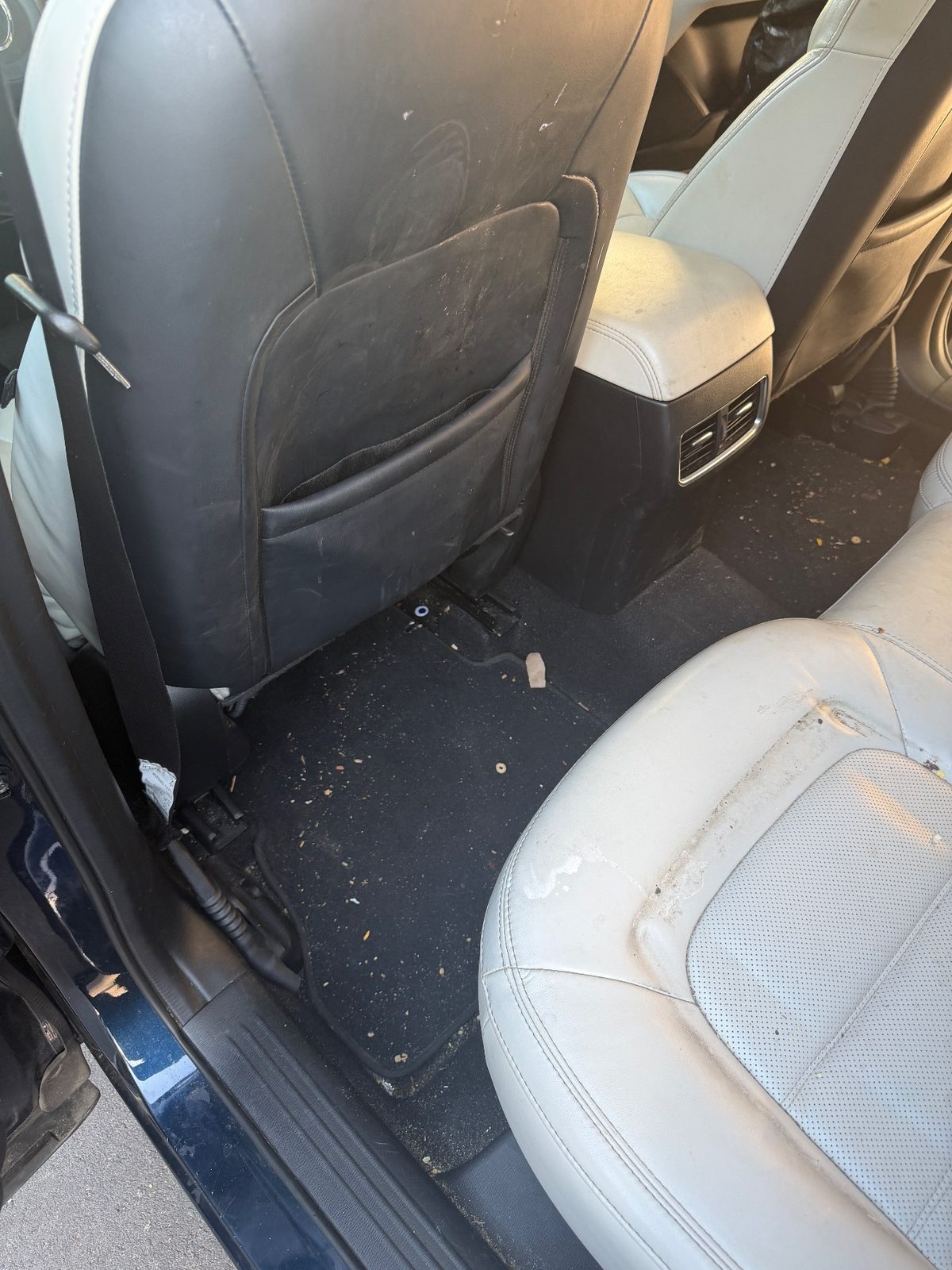 Rear footwell of a dark blue Mazda CX-5 before detailing, with crumbs on the carpet and scuffed seat-back leather
