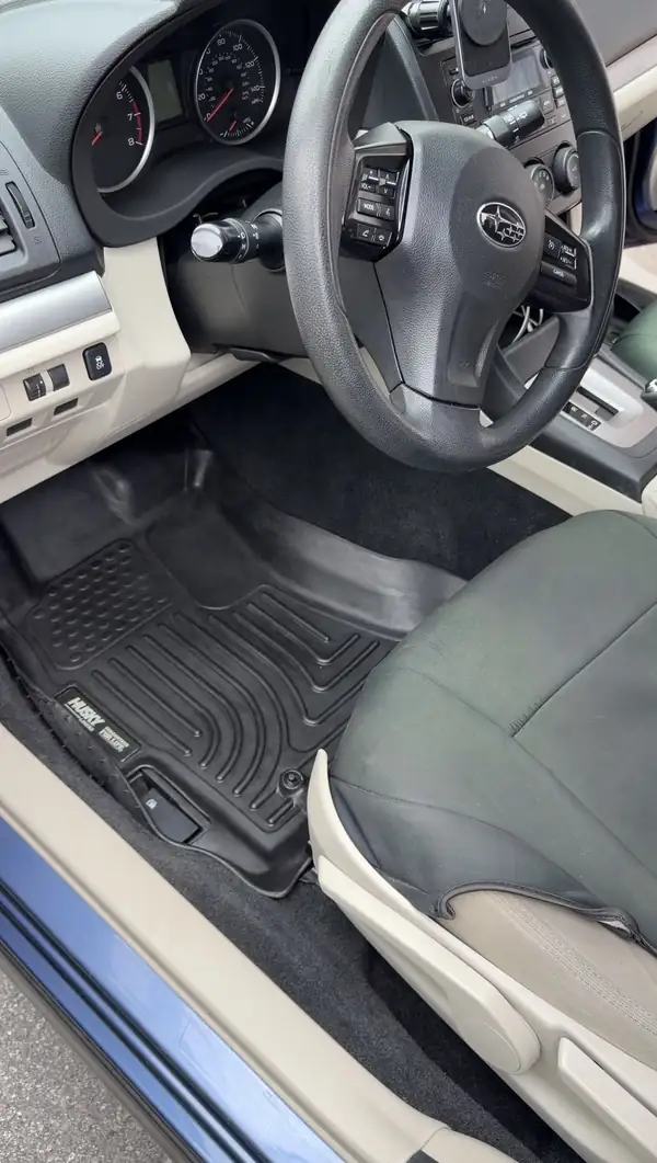 Clean driver footwell of a blue Subaru after detailing, the black rubber floor liner clear of dirt with the Subaru steering wheel above.