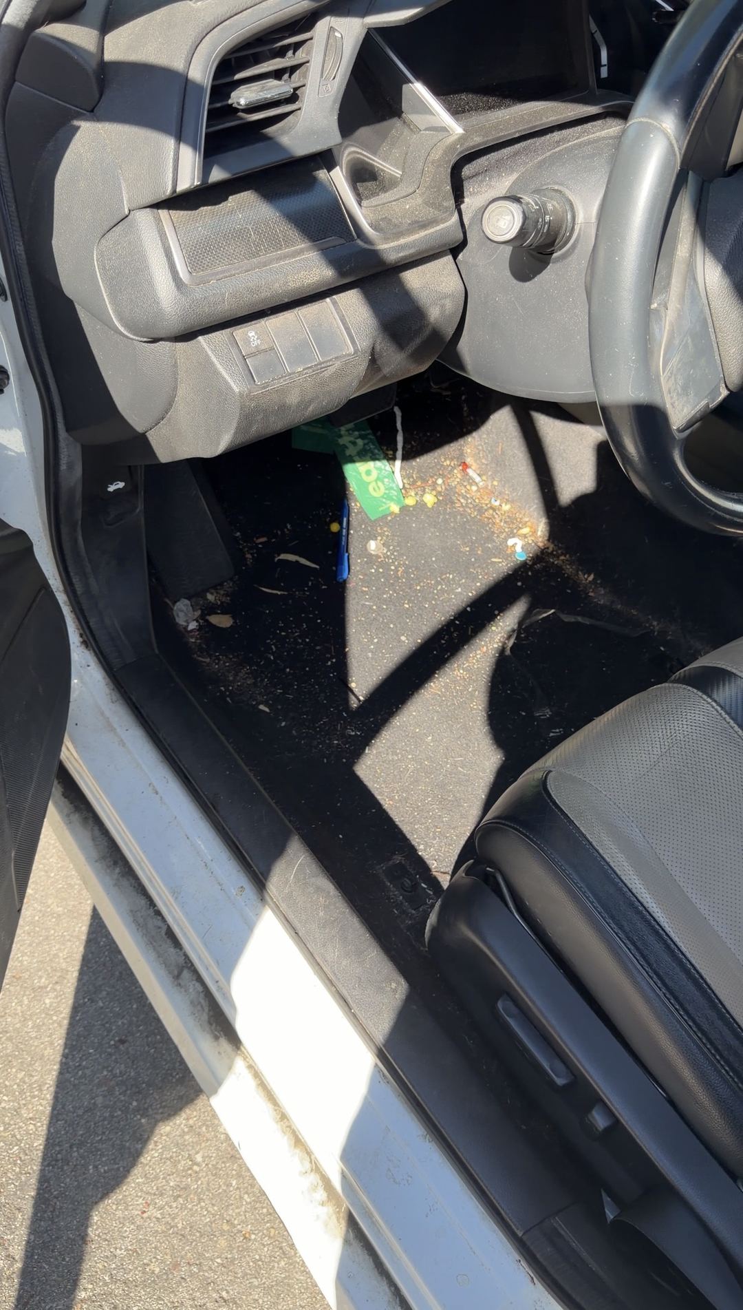 Driver footwell of a Honda Civic before cleaning, carpet strewn with candy debris, a wrapper and a pen