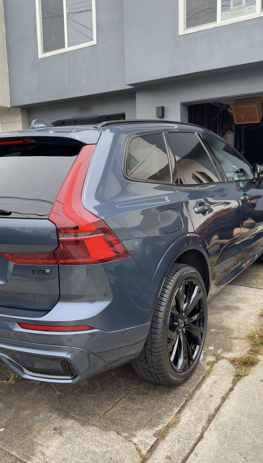 Blue-grey Volvo XC60 T8 from the rear corner, glossy paint and black wheels, parked by a house