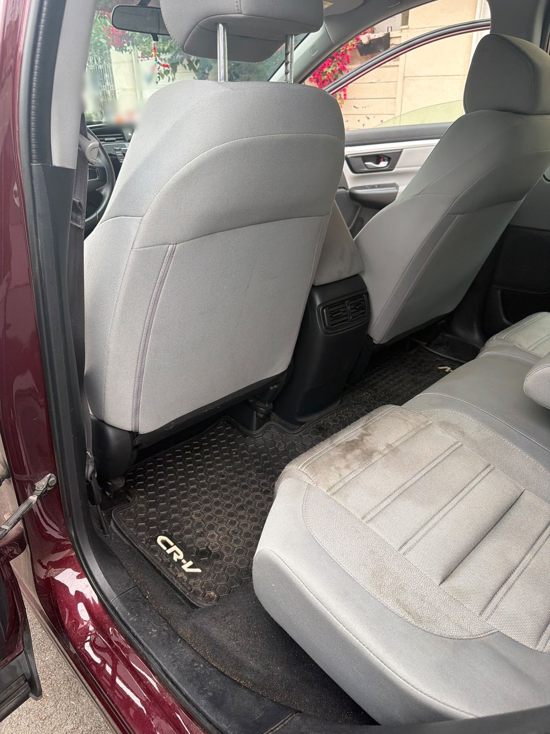 Rear footwell of a maroon Honda CR-V before detailing, a dusty rubber mat and a stained grey cloth seat cushion