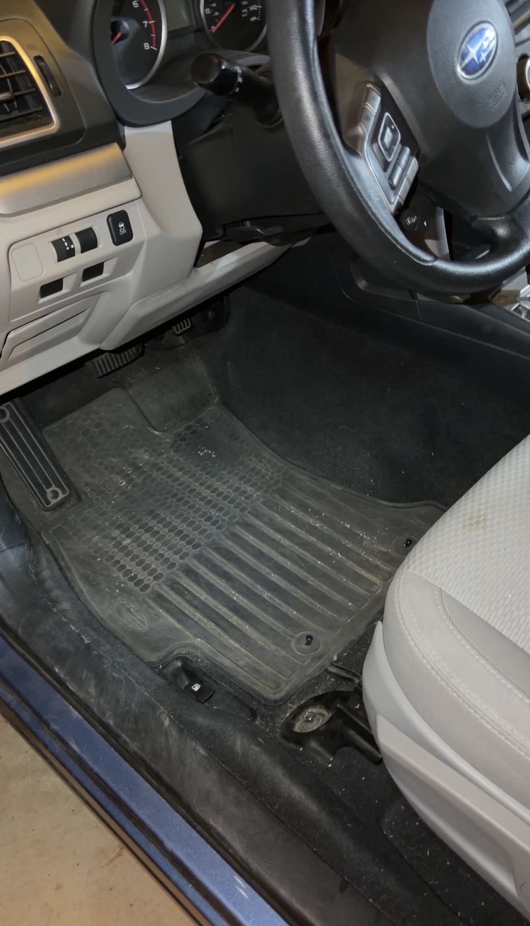 Driver's footwell of a blue Subaru Forester before detailing, a grey film of dust and grit over the black rubber all-weather mat.