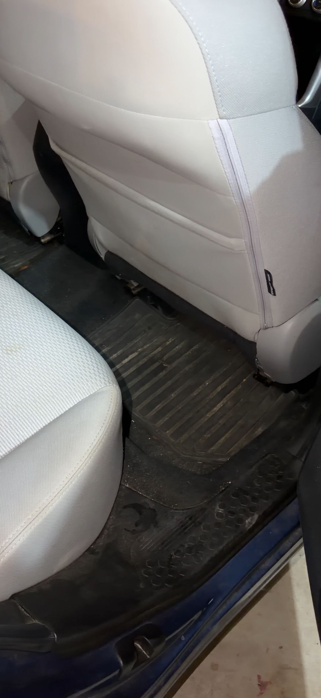 Rear passenger-side footwell of a blue Subaru Forester before detailing, grit and a twig caught in the ribs of the black rubber floor mat beside the light grey cloth seat.