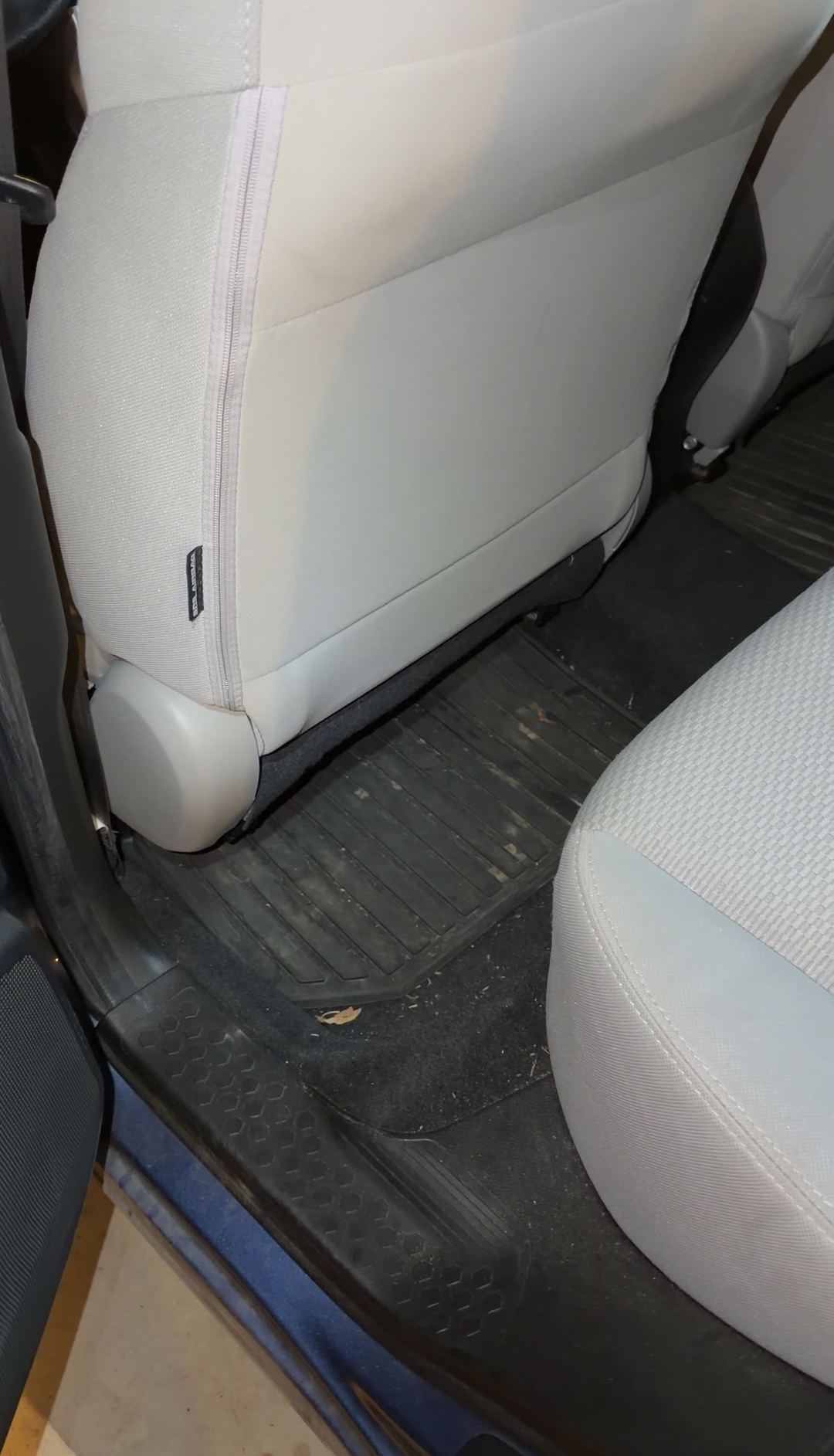 Rear driver-side footwell of a blue Subaru Forester before detailing, dried leaf fragments and grit on the black rubber mat and carpet.