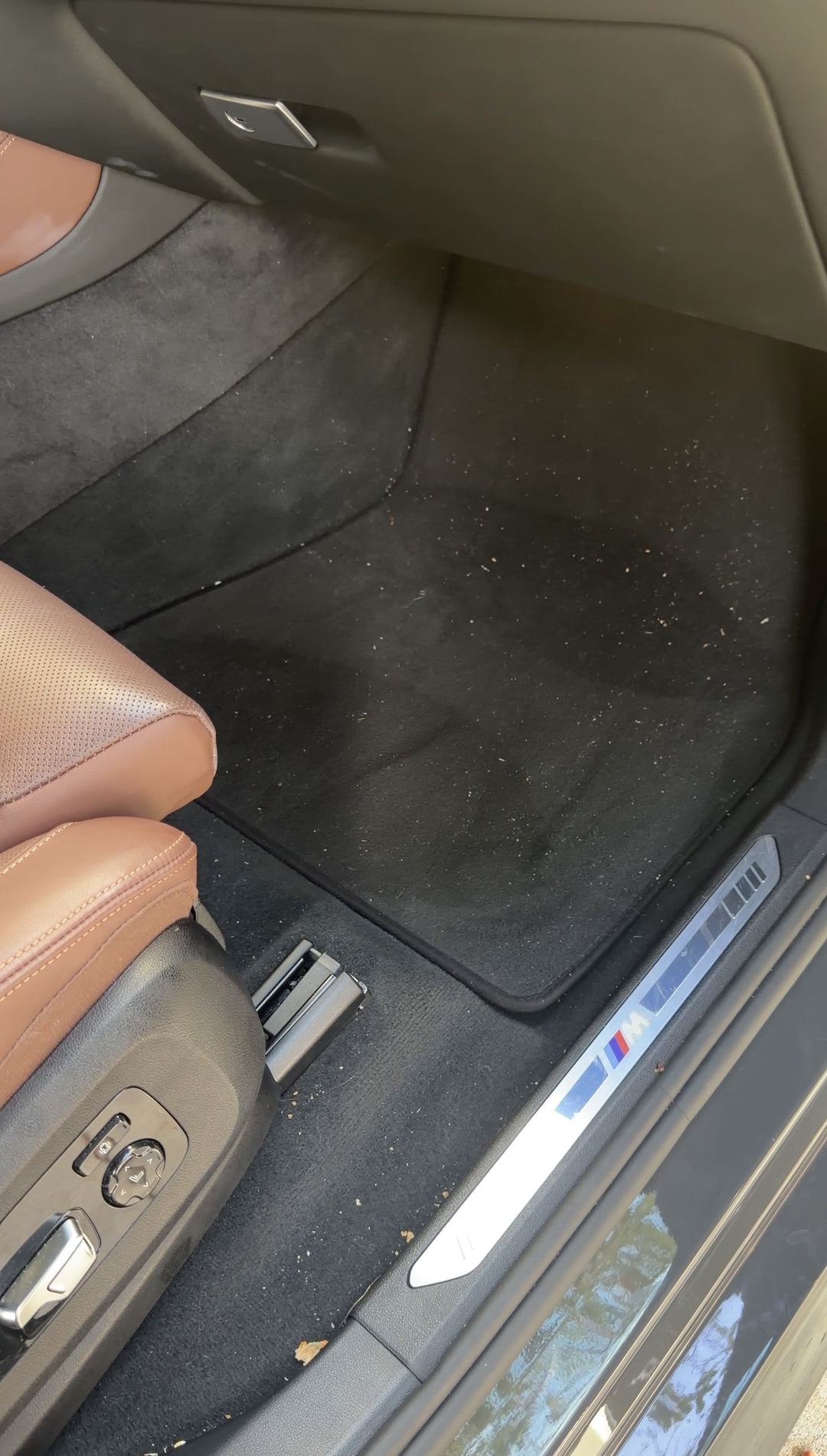 Front passenger footwell of a BMW X5 before vacuuming, with lint and grit across the black floor mat