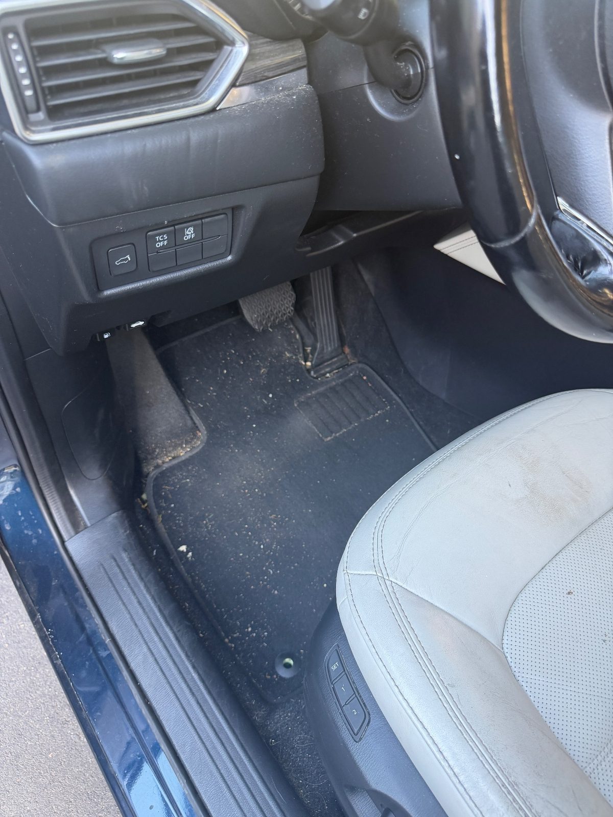 Driver footwell of a dark blue Mazda CX-5 before detailing, mat covered in crumbs and debris