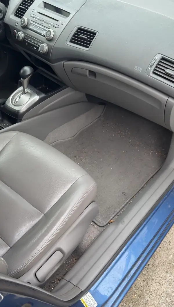 Front passenger footwell of a Honda Civic before cleaning, carpet mat covered in crumbs and leaf debris with grit along the door sill.