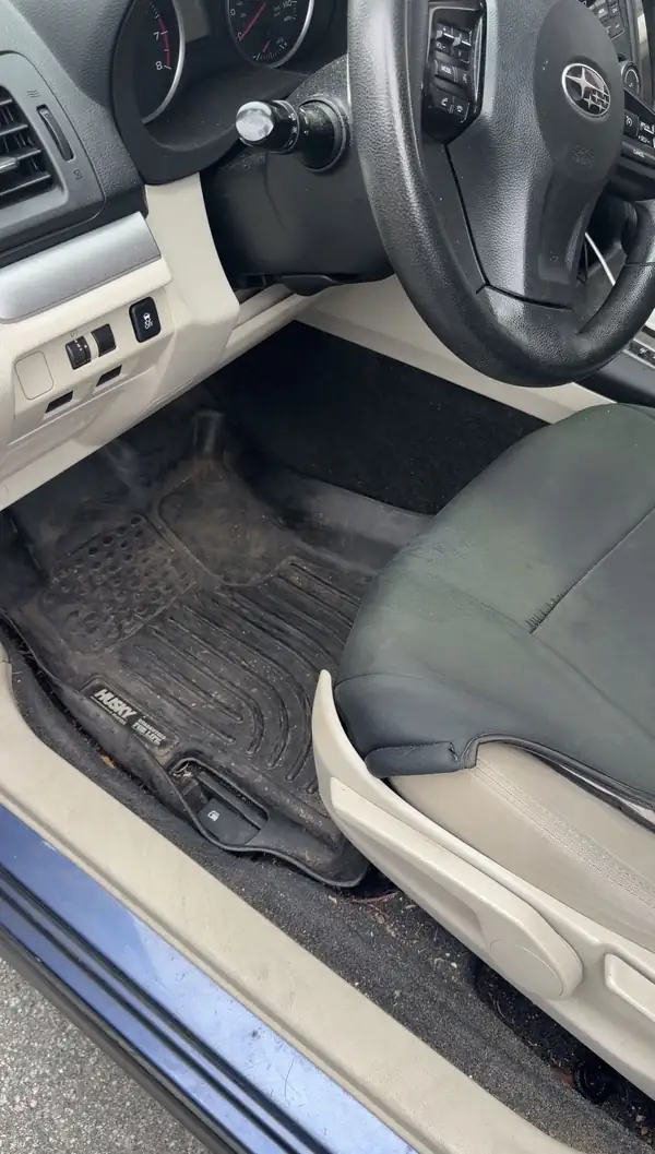 Driver footwell of a blue Subaru before cleaning, the black rubber floor liner grey with dust and scattered with crumbs and pine needles.