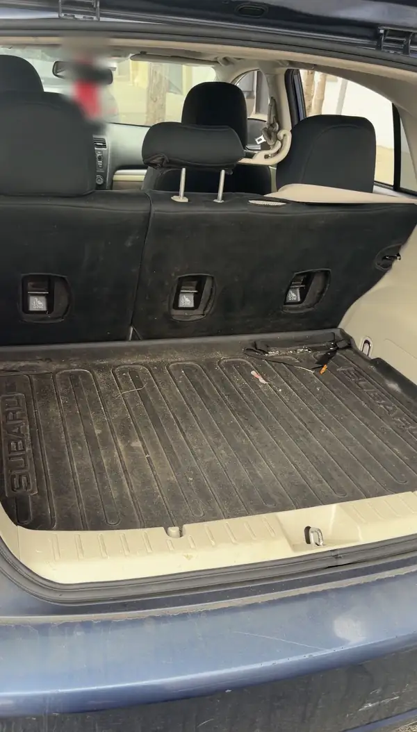 Cargo area of a blue Subaru hatchback before cleaning, the ribbed Subaru load tray dulled by dust with crumbs and plant debris in the grooves.