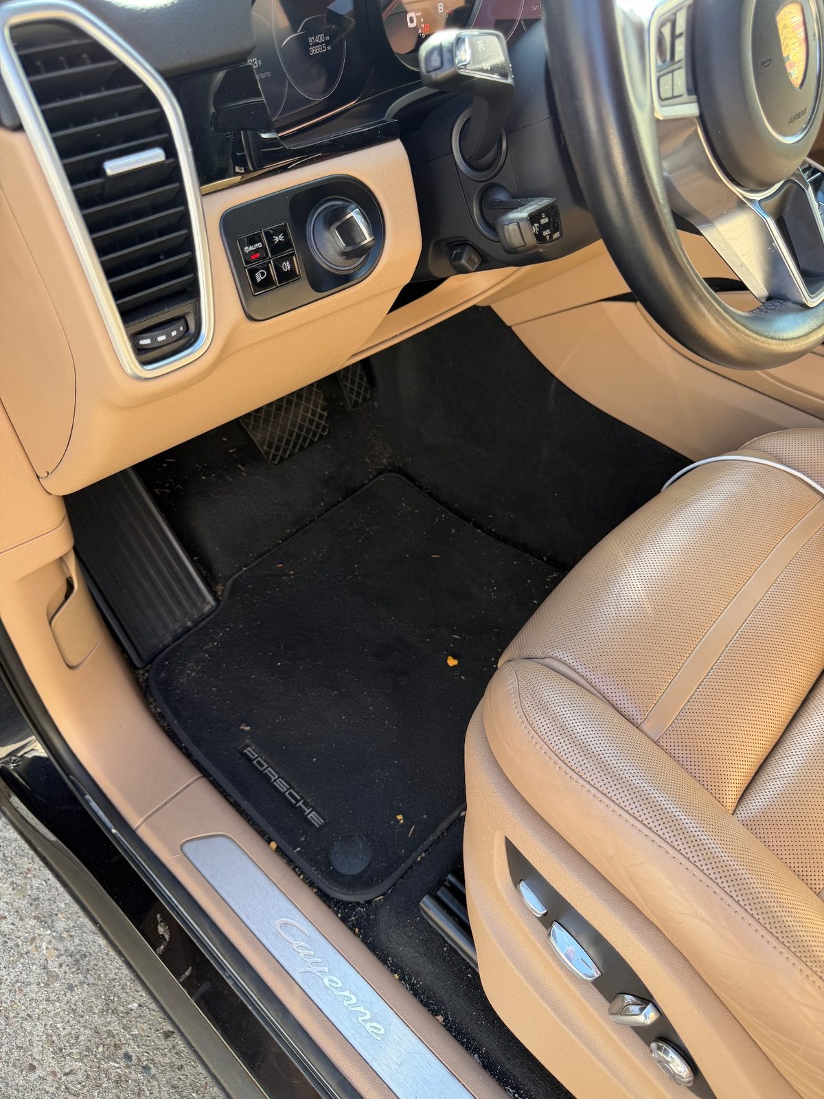 Driver footwell of a dark blue Porsche Cayenne before vacuuming, carpet mat covered in lint and crumbs