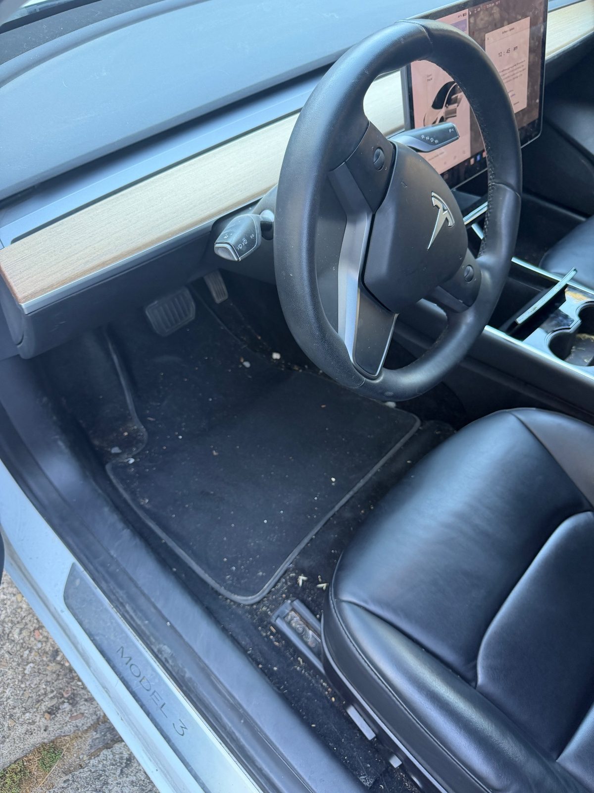 Driver footwell of a white Tesla Model 3 before detailing, with crumbs and grit on the floor mat
