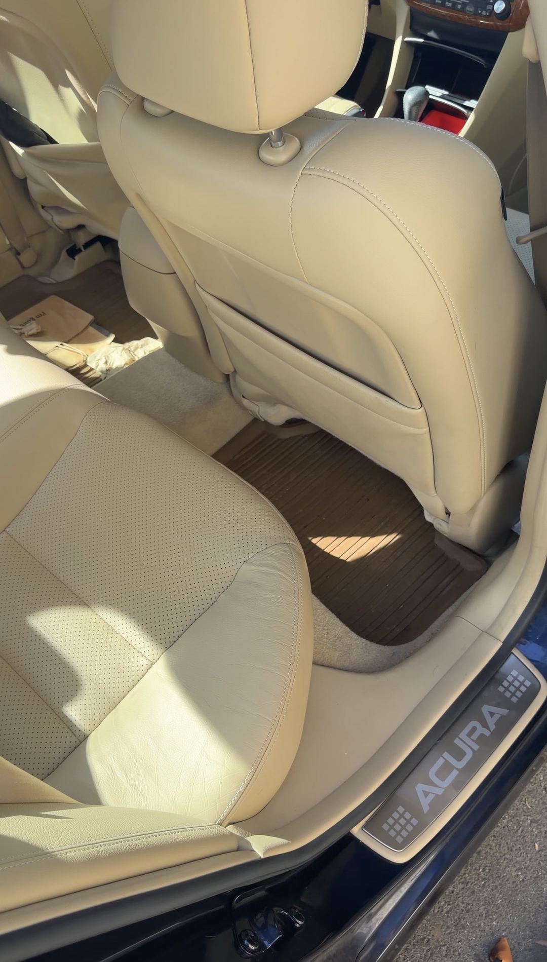Rear passenger-side footwell of a tan-leather Acura sedan before detailing, with dust and grit on the rubber floor mat.