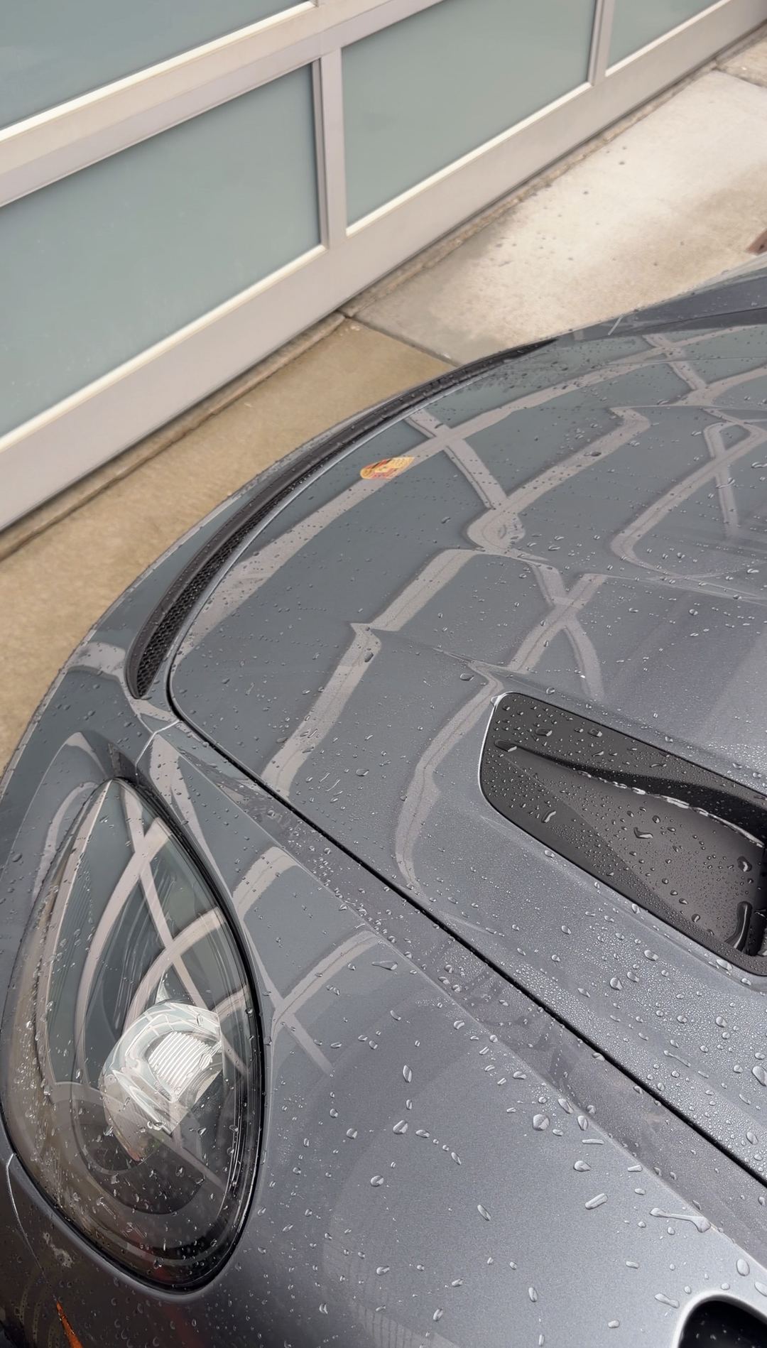 Rinse water beaded across the bonnet and headlight of a grey Porsche 718 Spyder RS.