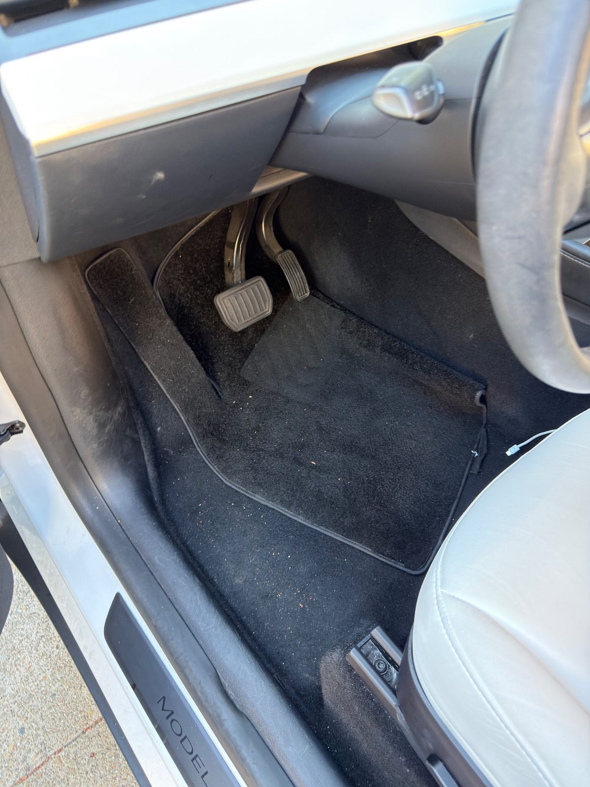 Driver footwell of a white Tesla Model Y before detailing with crumbs and specks on the black carpet