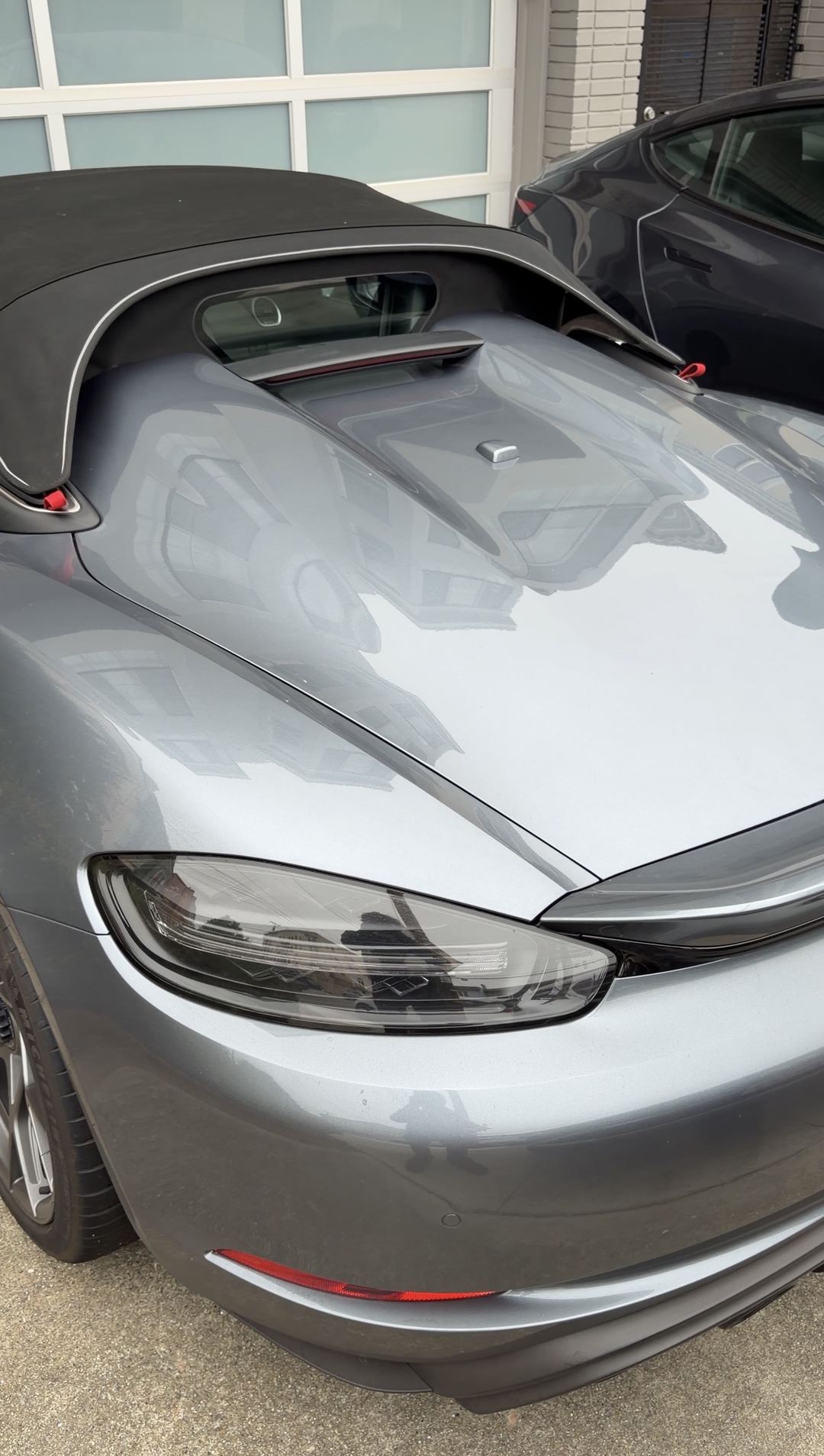 Grey Porsche 718 Spyder RS seen from the rear three-quarter with its black soft top raised, parked in front of a frosted-glass garage door before a mobile detailing wash.