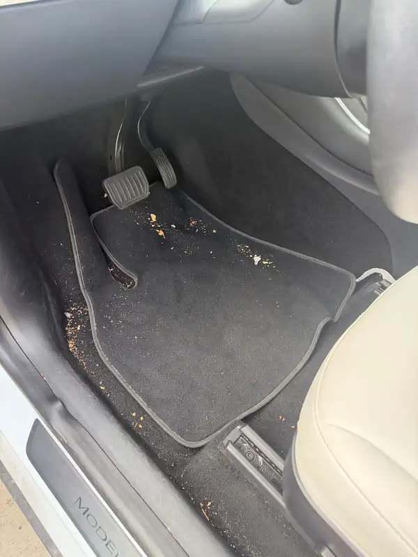 Driver footwell of a white Tesla Model Y before detailing, with crumbs and debris on the carpet mat