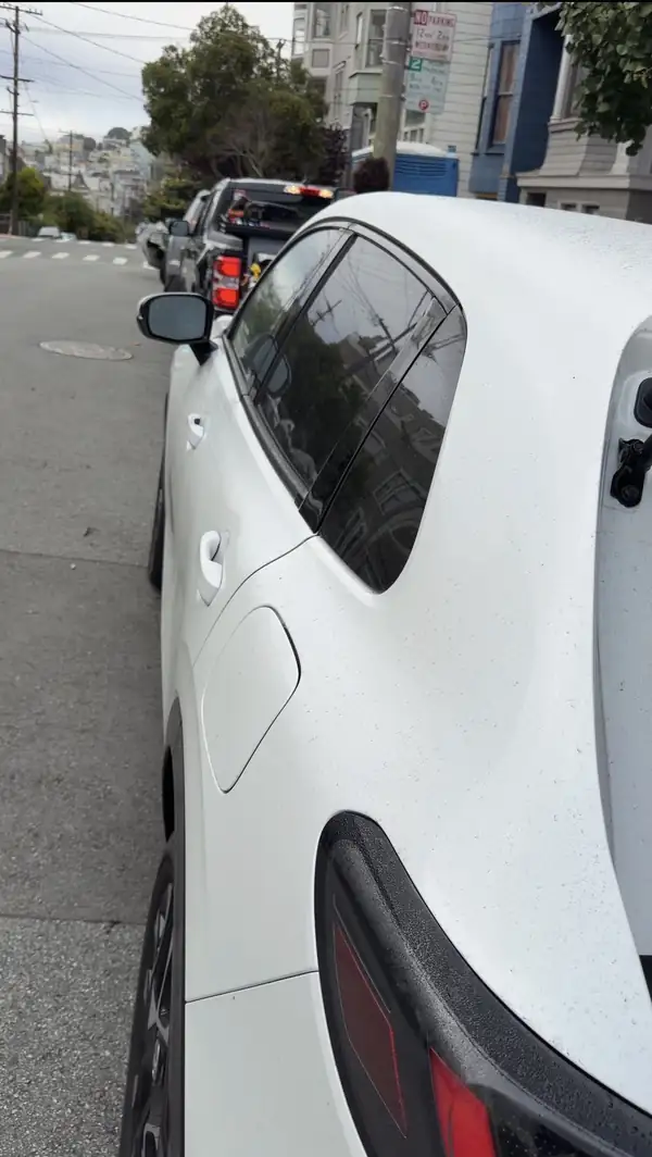 Rear three-quarter view of a white Honda HR-V parked on a hilly city street, its paint covered in a fine layer of road grime before washing.