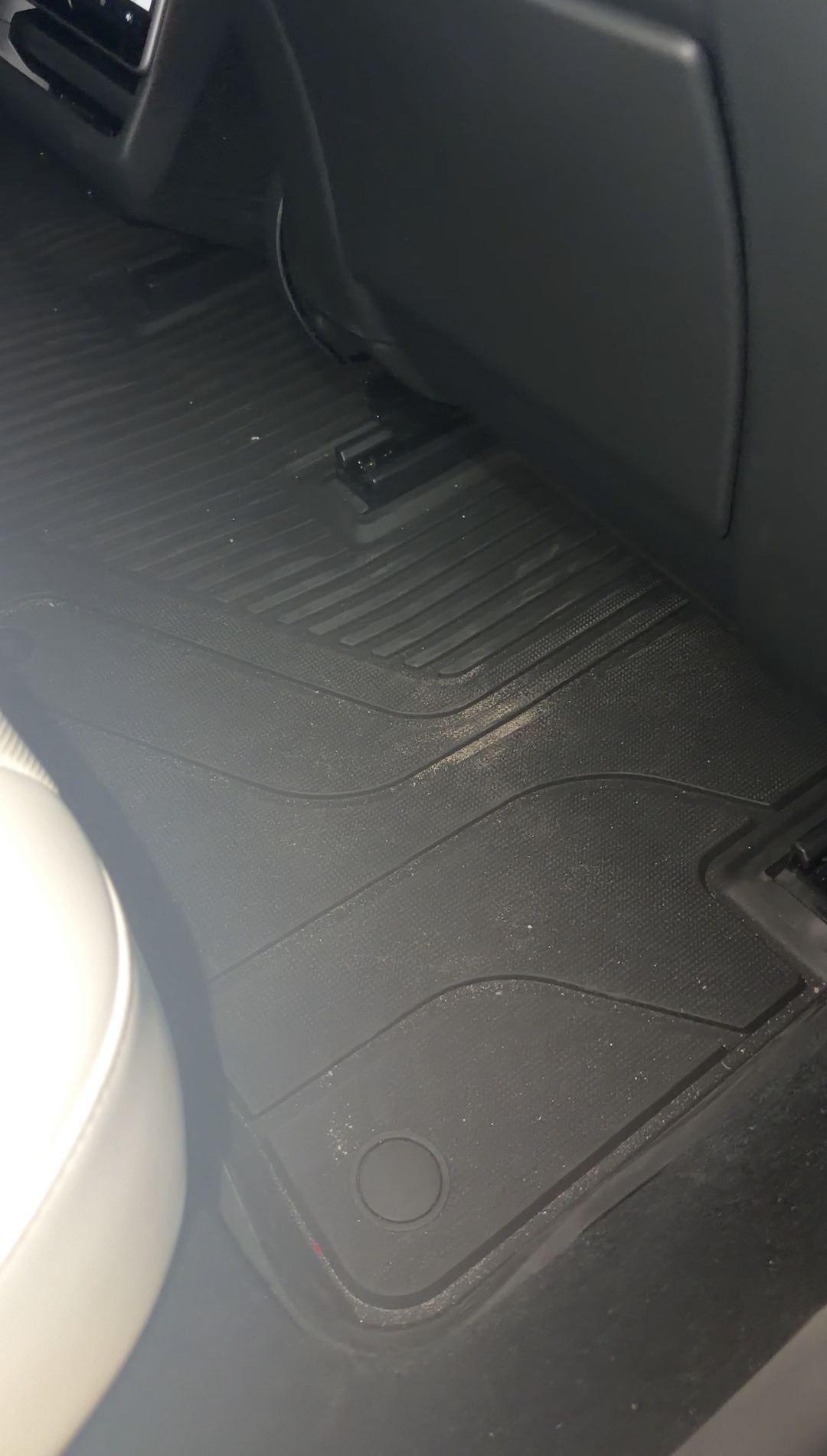 Rear passenger footwell of a Tesla Model 3 before detailing, with sandy grit scattered across the black all-weather floor mat