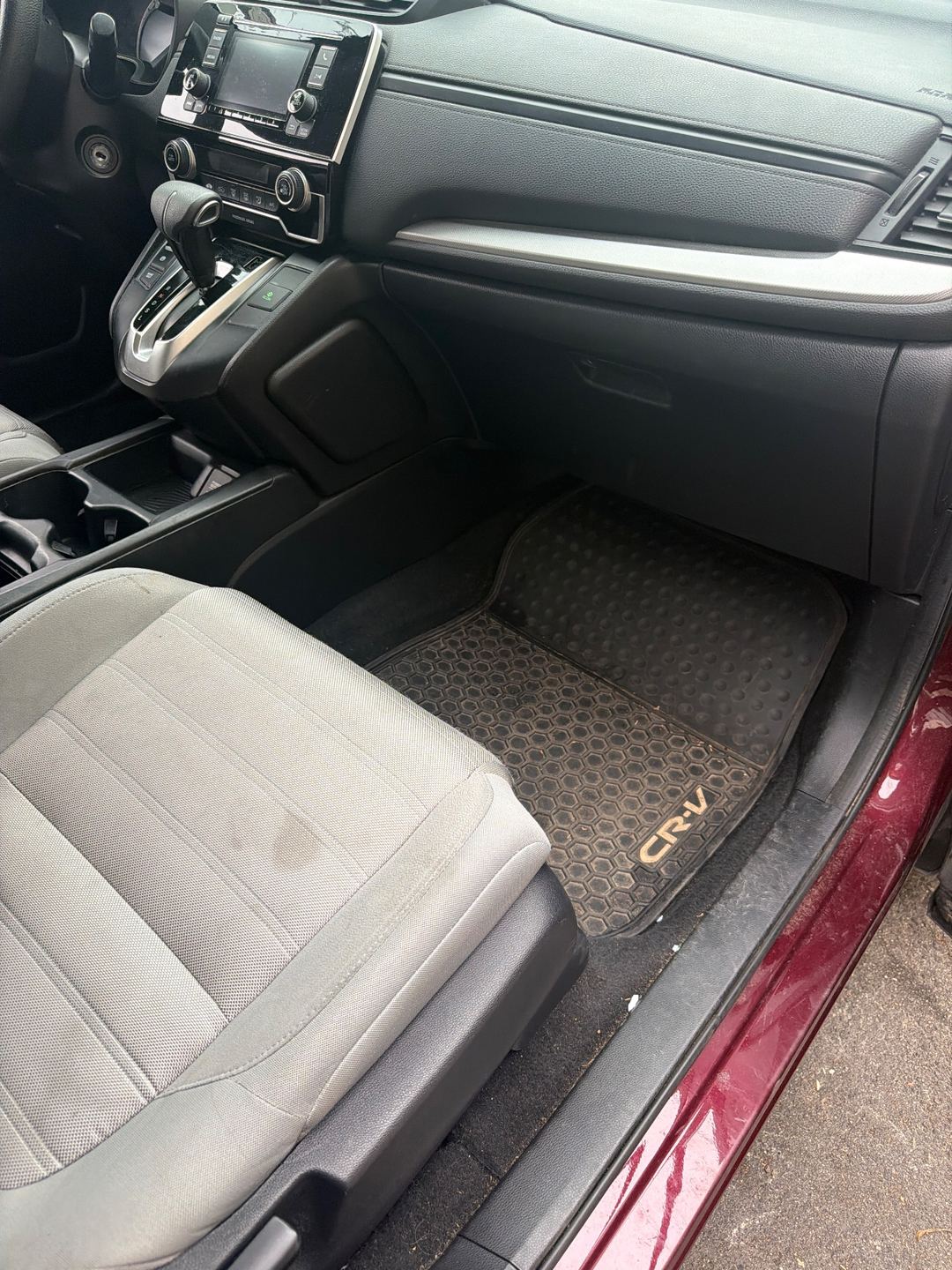 Front passenger footwell of a maroon Honda CR-V before detailing, the rubber mat dusty and a stain on the cloth seat