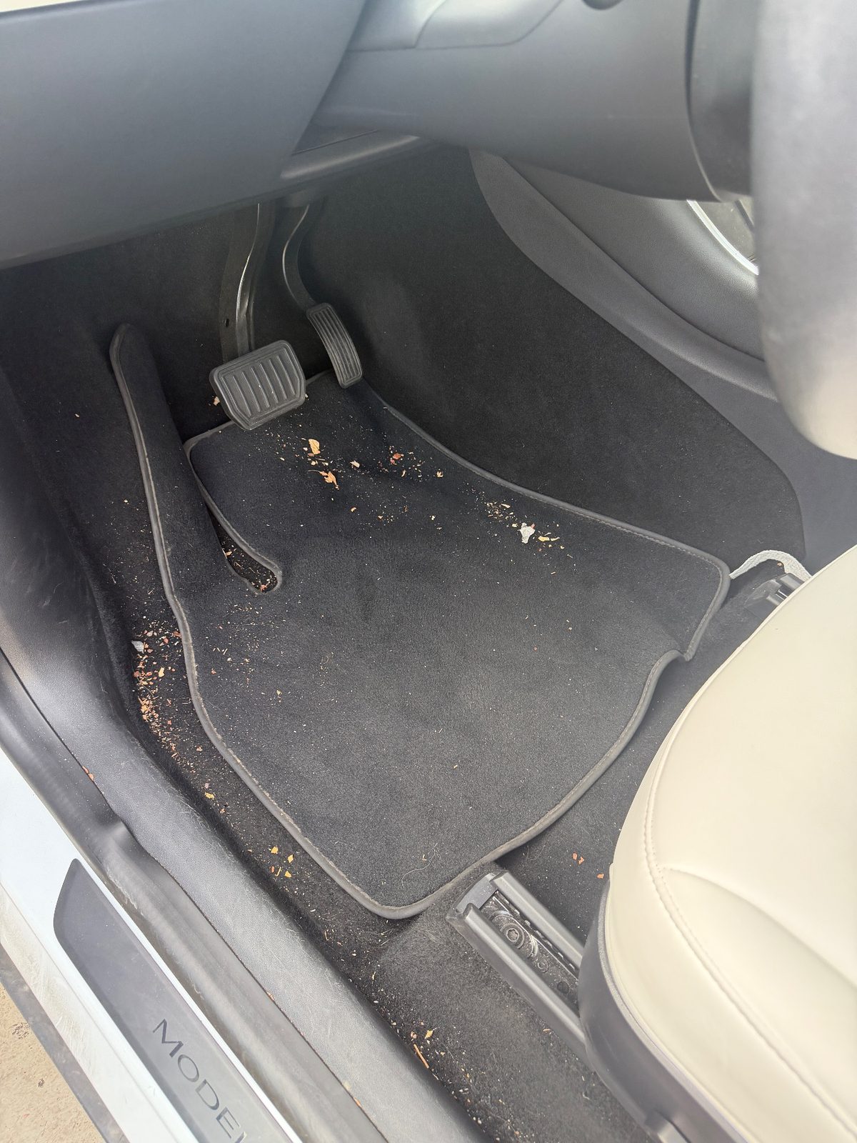 Driver footwell of a white Tesla Model Y before detailing, with crumbs and debris on the carpet mat
