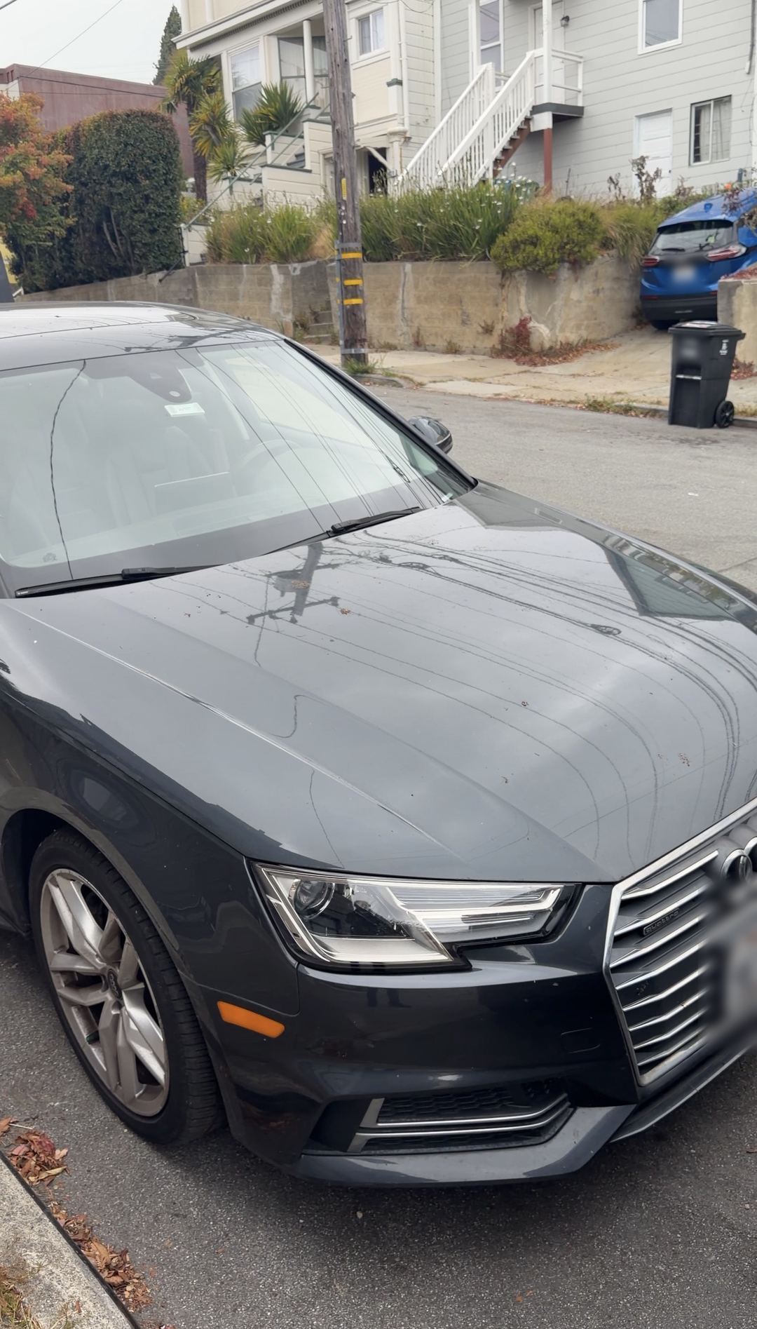 Grey Audi A4 with streaks and leaf bits on the hood, parked on a hillside residential street