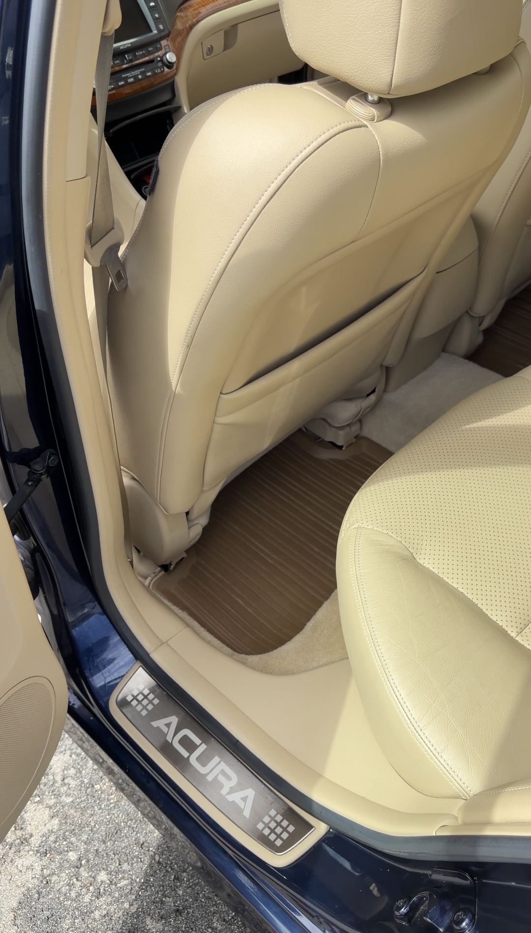 Cleaned rear driver-side footwell of a tan-leather Acura sedan after detailing, with a clean rubber mat and bright carpet at the door sill.