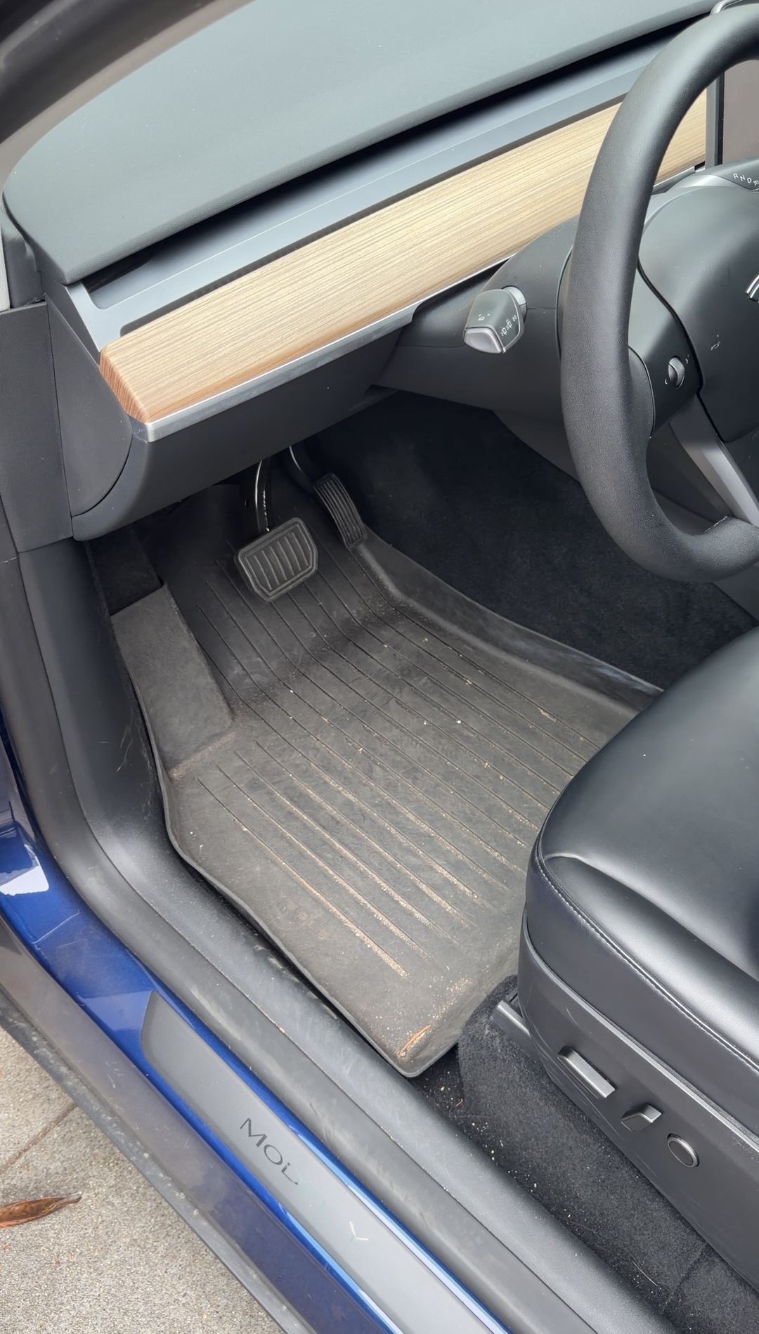 Driver's footwell of a blue Tesla Model Y before detailing, the rubber floor liner dulled by dust with grit packed into its channels.
