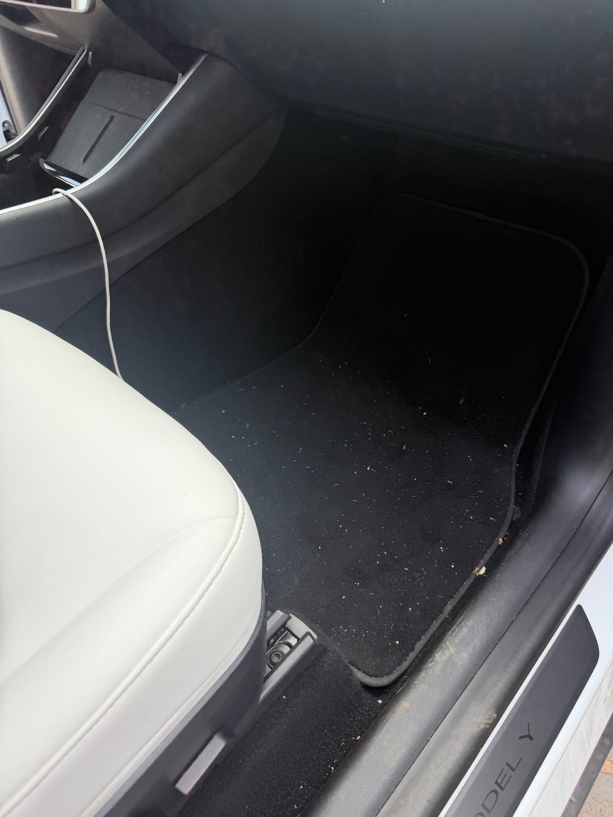 Passenger footwell of a white Tesla Model Y before detailing, with lint and dust on the carpet mat