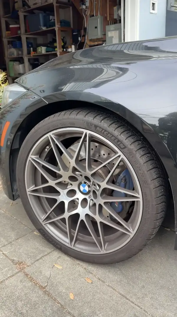 Forged star-spoke wheel with a blue M brake caliper on a black BMW M3 at a garage doorway