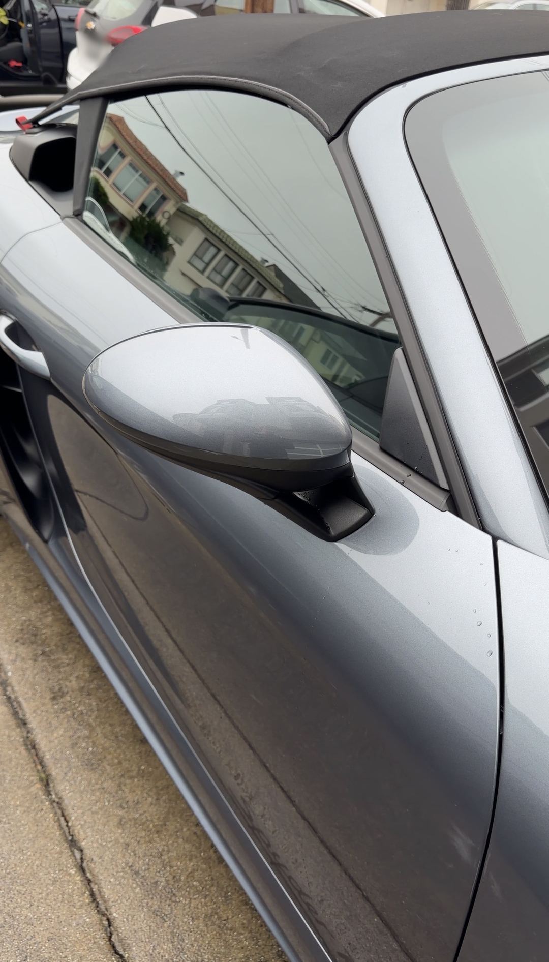 Door mirror and A-pillar of a clean grey Porsche 718 Spyder RS with its soft top raised.