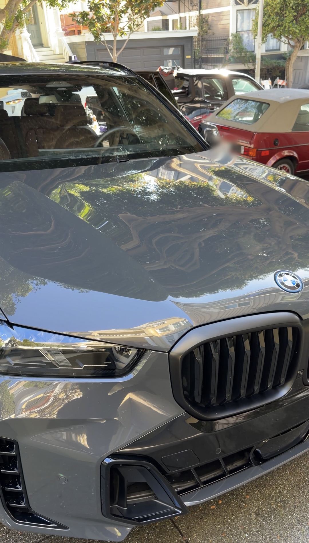 Front three-quarter of a dark grey BMW X5 after detailing, its hood mirroring the trees and houses above