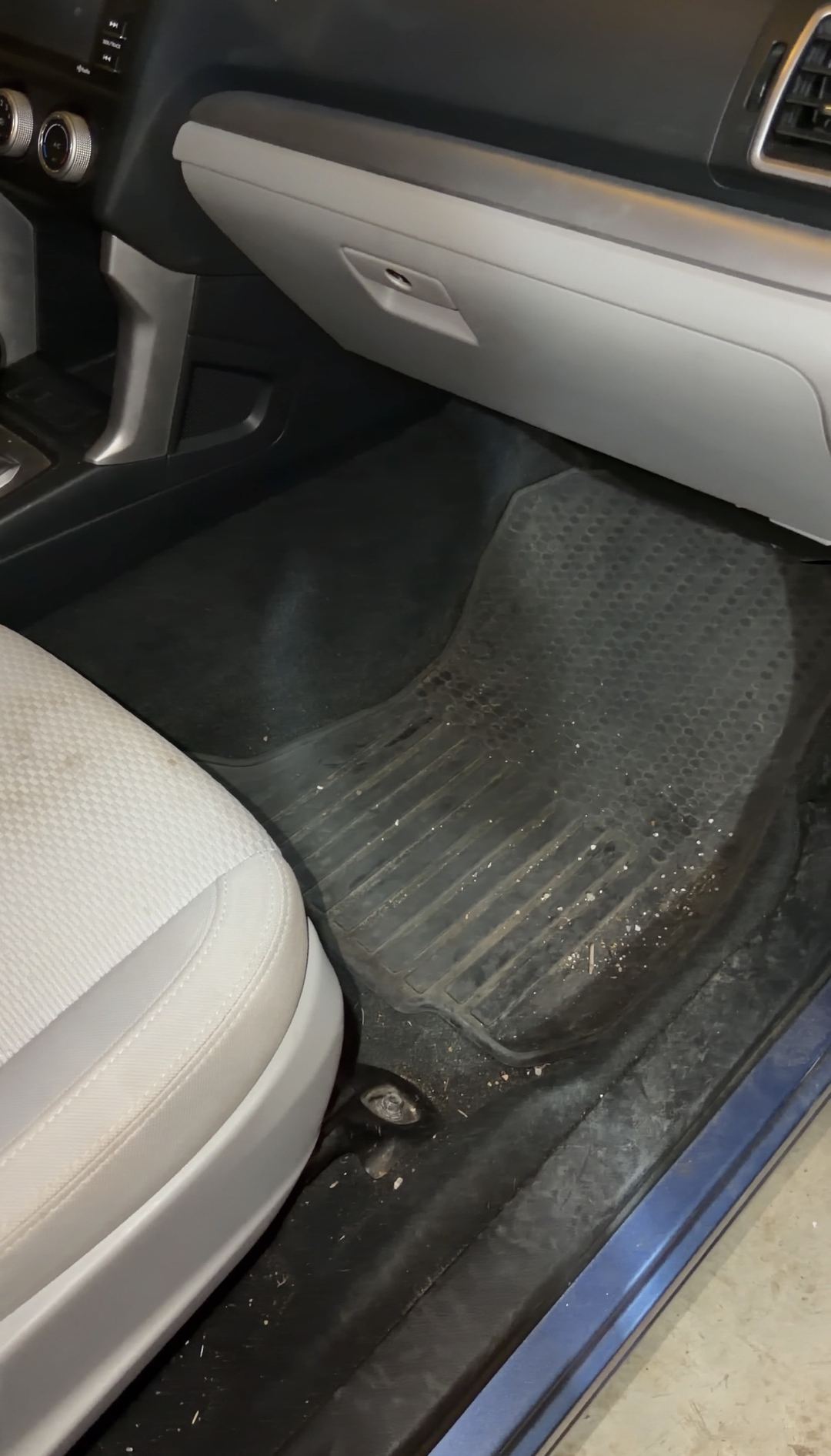 Front passenger footwell of a blue Subaru Forester before detailing, crumbs and grit scattered across the black rubber all-weather mat below the grey glovebox.