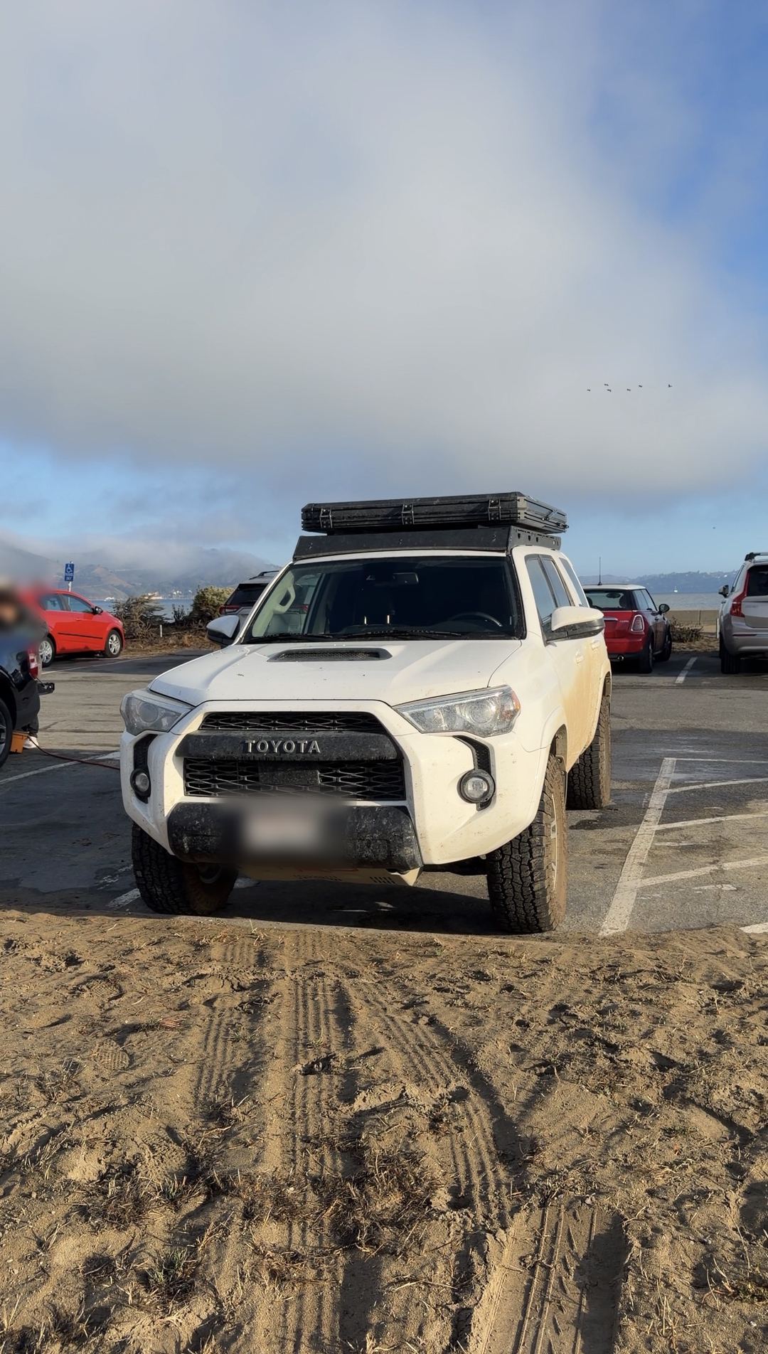 White Toyota 4Runner TRD Pro with a rooftop tent parked beside sand under a foggy sky, before cleaning