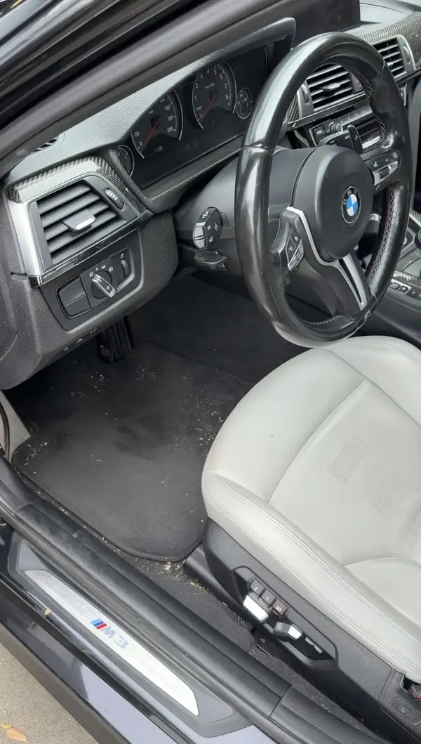Driver's footwell of a BMW M3 before cleaning, with grit and leaf debris on the black floor mat beside the lit M3 door sill