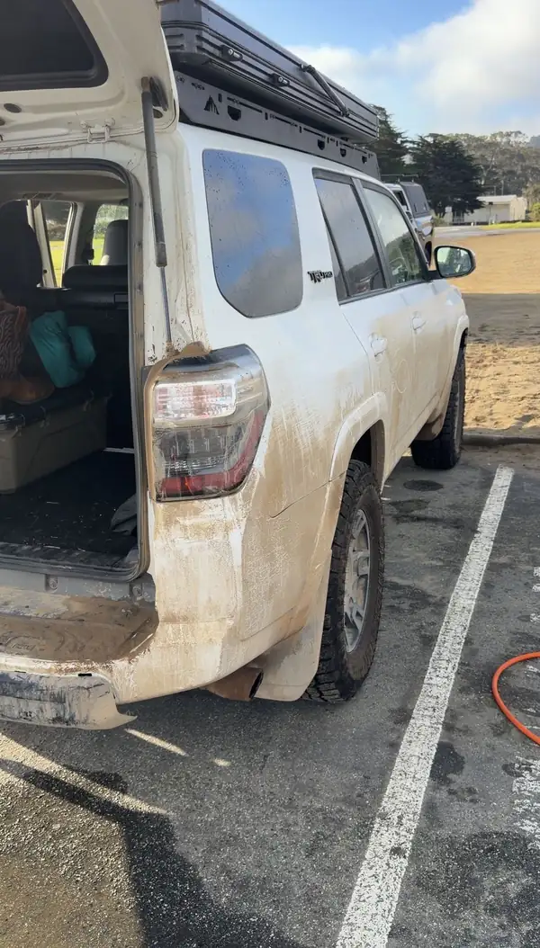 Passenger-side rear of a white Toyota 4Runner with mud-streaked panels, open tailgate and rooftop tent