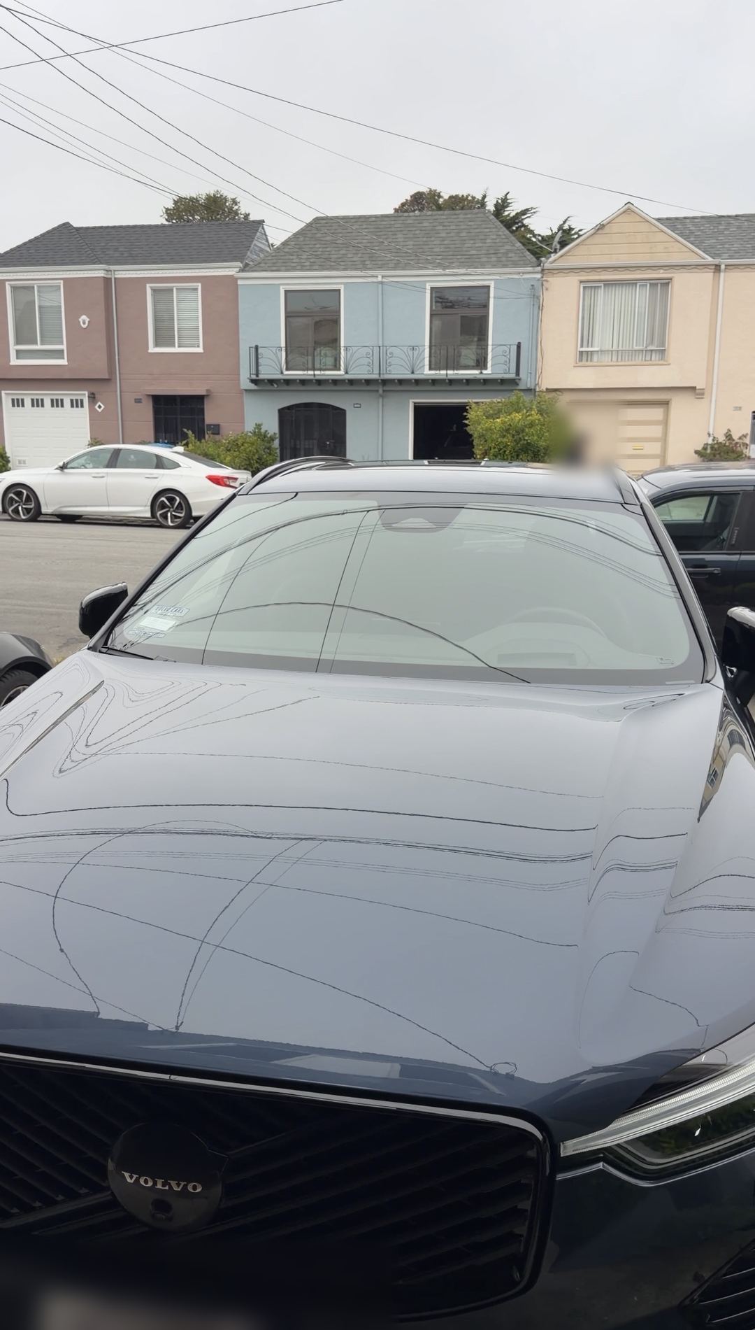 Hood of a blue-grey Volvo XC60 with thin liquid lines on the paint, houses across the street