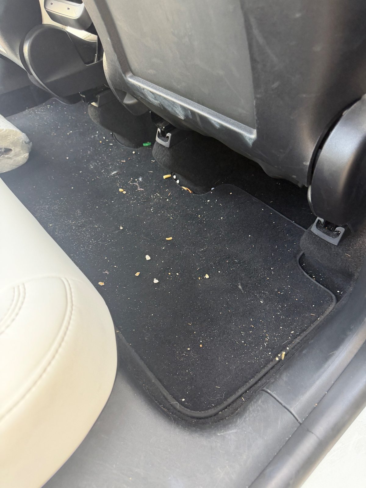 Rear footwell of a white Tesla Model Y before detailing, with crumbs and lint on the carpet mat