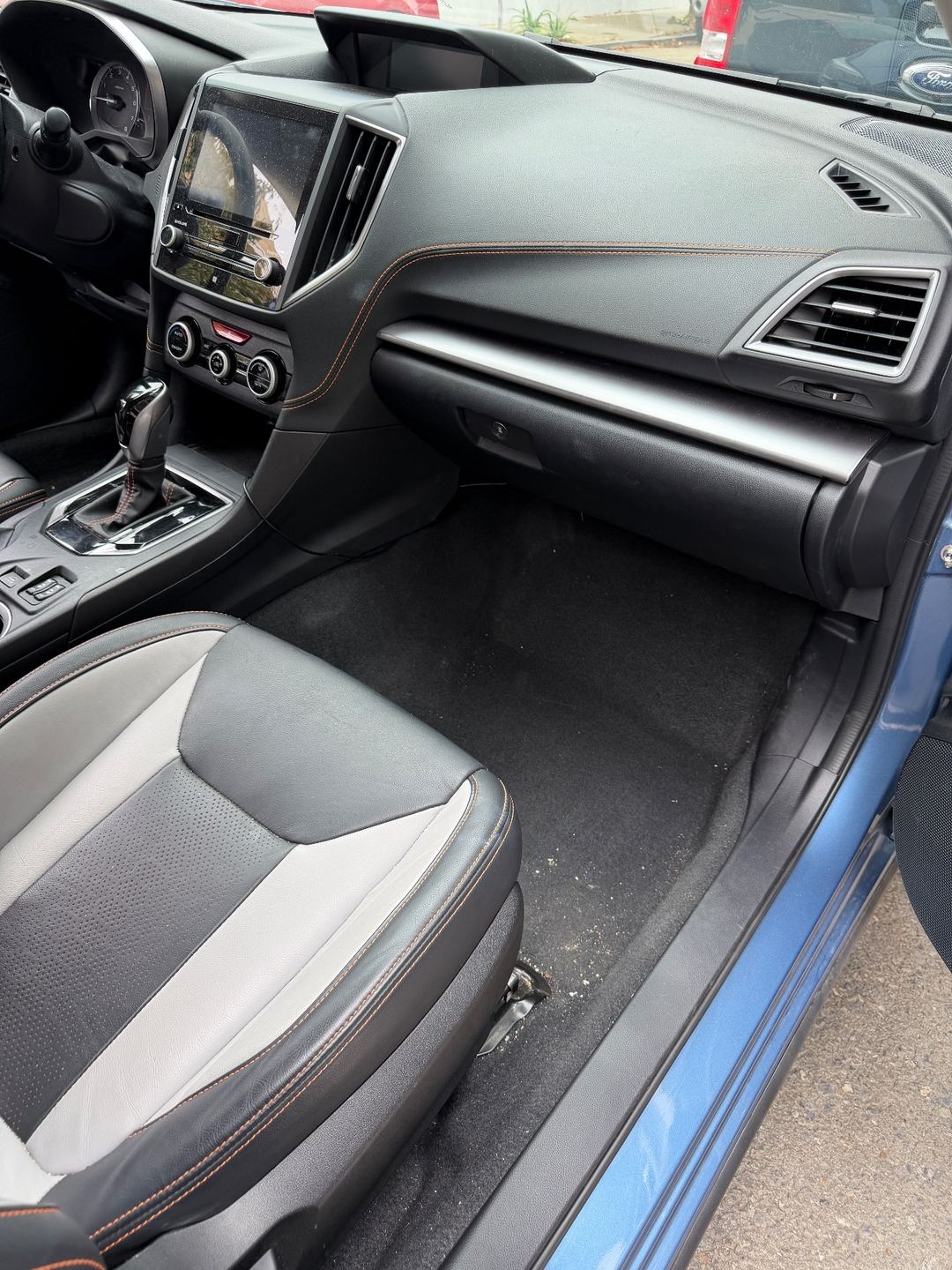 Front passenger footwell of a blue Subaru Crosstrek before detailing, with crumbs and grit on the carpet