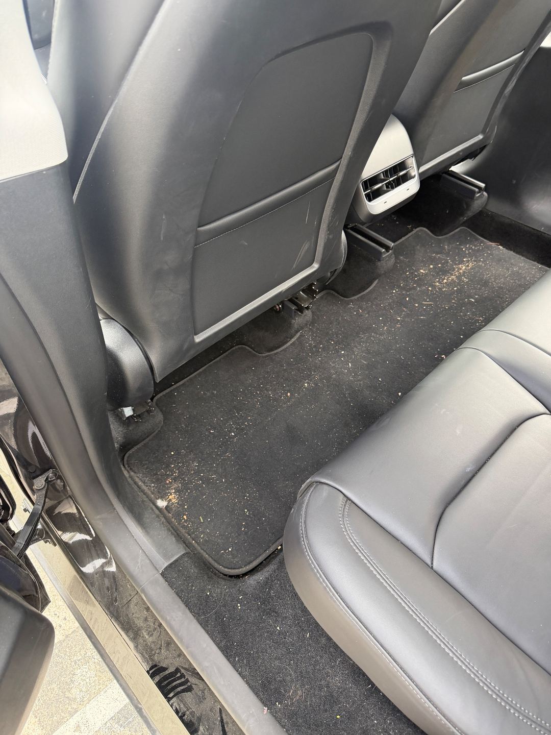 Rear footwell of a black Tesla Model Y before detailing, with sand and crumbs on the floor mat