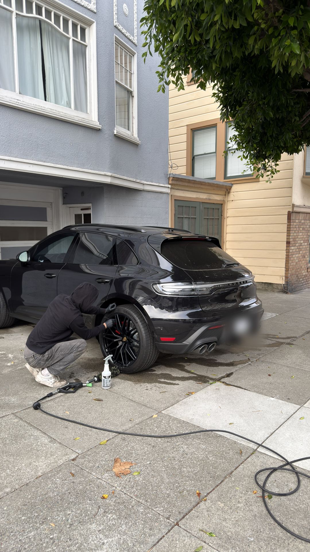 SF City Wash detailer kneeling beside a black Porsche Macan GTS on a San Francisco driveway, hand-cleaning the rear wheel.