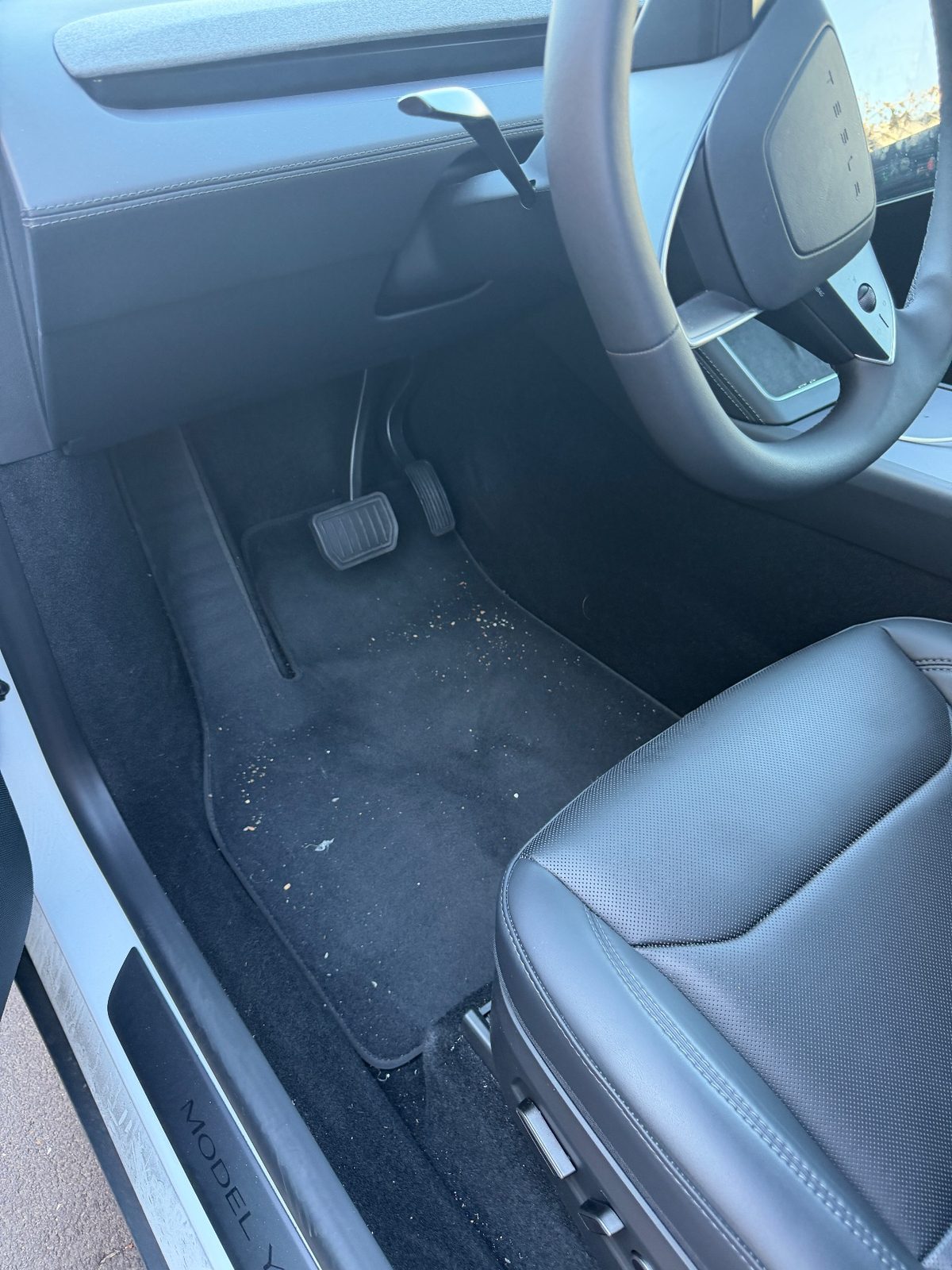 Driver footwell of a white Tesla Model Y before detailing, crumbs and grit on the black mat