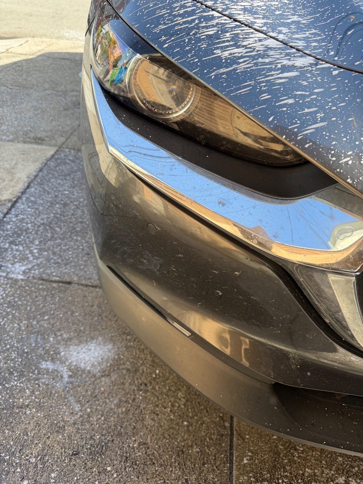 Front corner of a grey Mazda CX-30 before the wash, bumper dotted with bugs and grime
