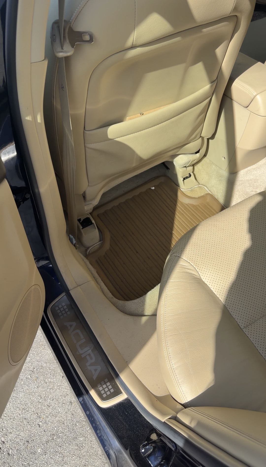 Rear driver-side footwell of a tan-leather Acura sedan before detailing, with grit on the rubber floor mat and a soiled carpet edge at the door sill.