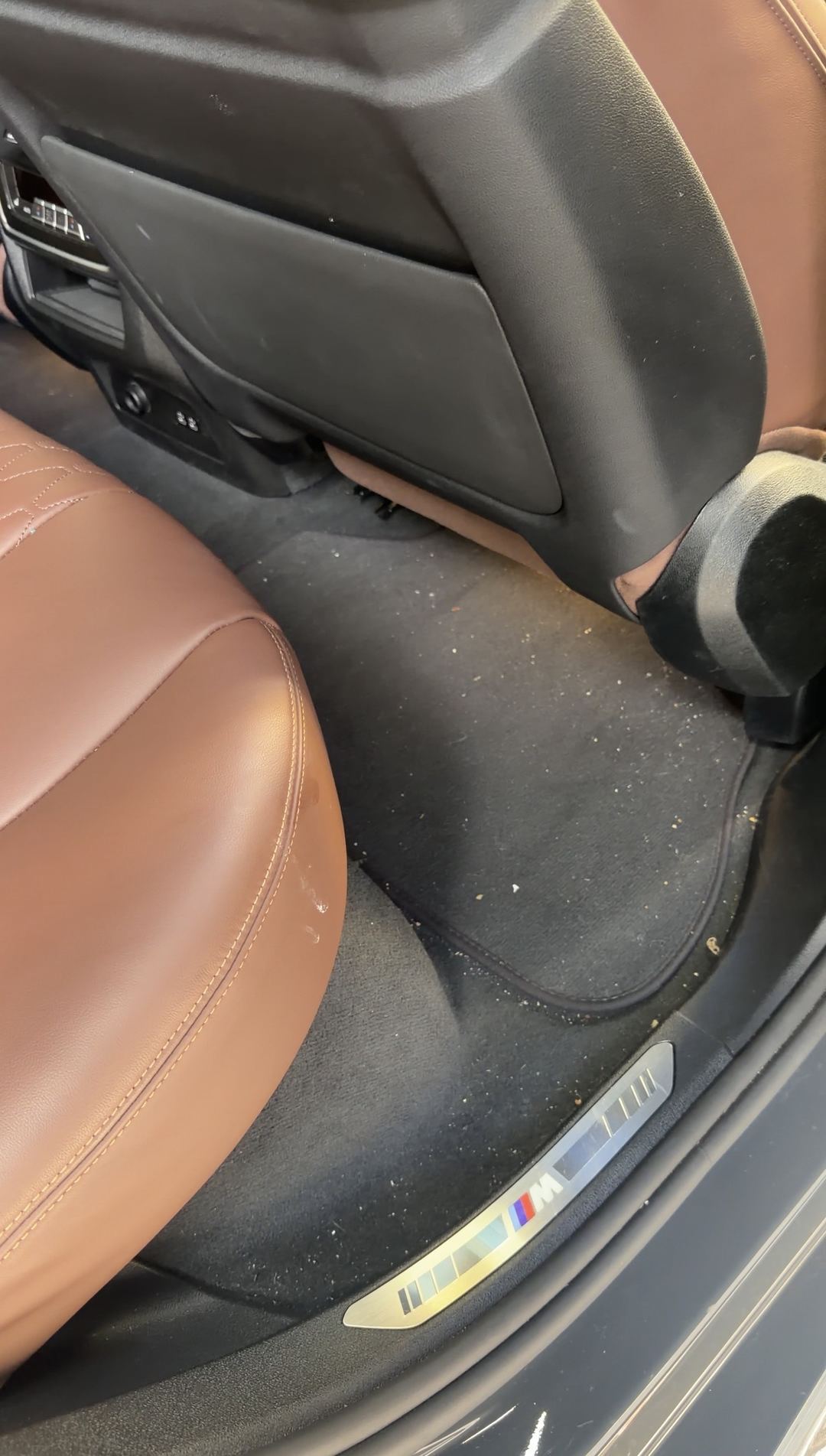 Rear passenger footwell of a BMW X5 before vacuuming, with crumbs scattered over the floor mat