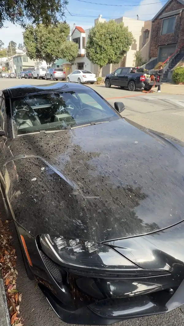 Hood of a black Toyota GR Supra before washing, covered in bird droppings, water spots and road film on a San Francisco street.