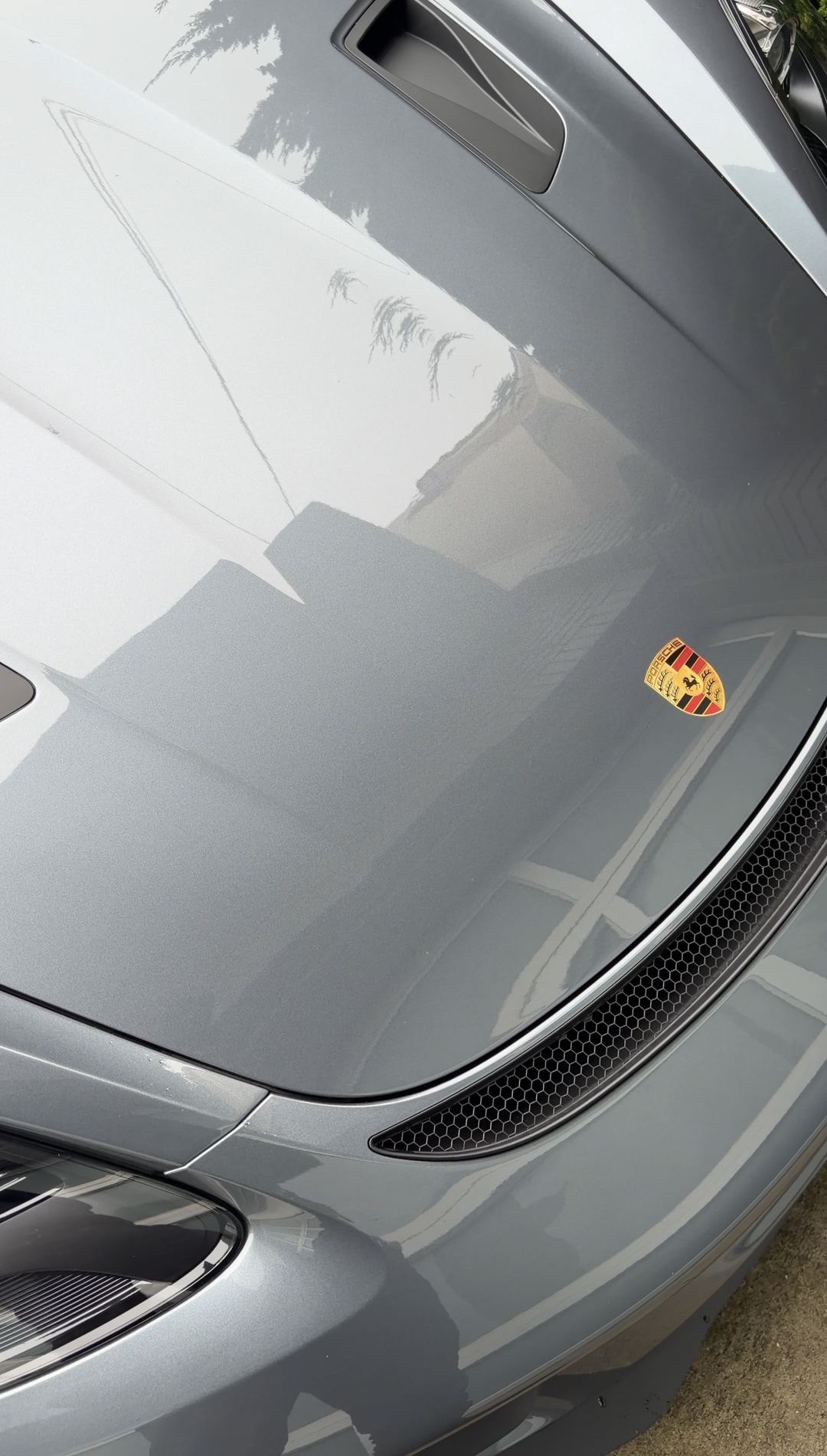 The bonnet of a grey Porsche 718 Spyder RS beside the Porsche crest, clean after the bird dropping was removed.