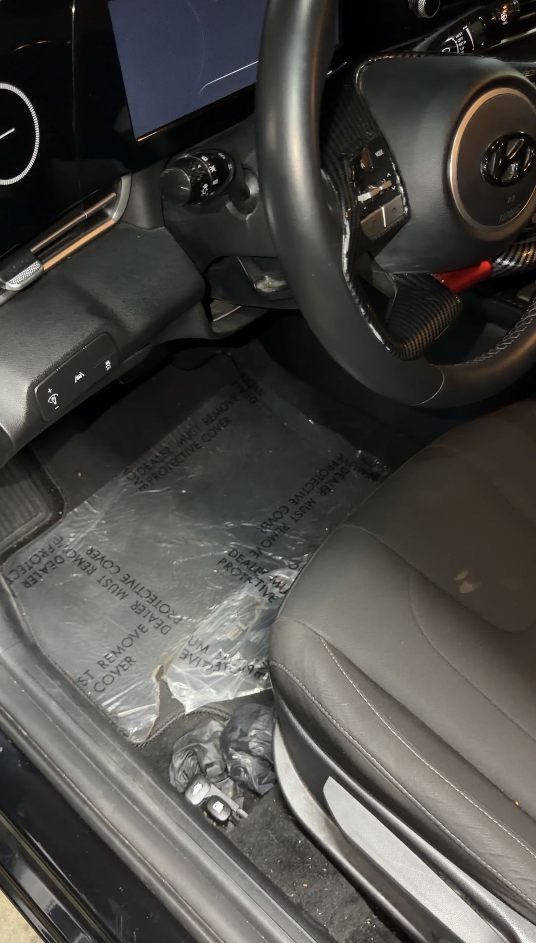 Driver footwell of a black Hyundai Elantra before cleaning, with debris by the seat and dealer plastic over the mat