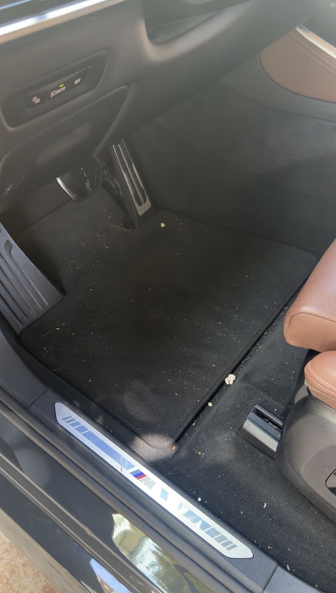 Driver footwell of a BMW X5 before vacuuming, with grit and crumbs covering the floor mat