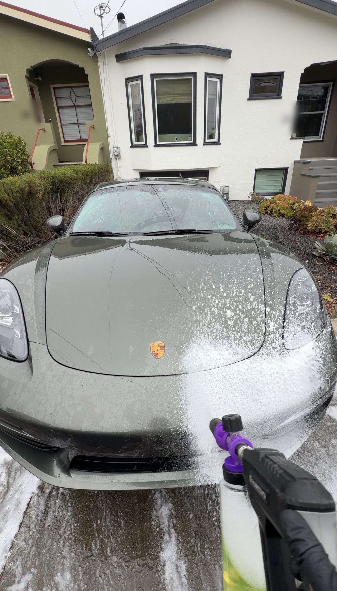 Snow foam being applied to the nose of a green Porsche 718 Cayman S with a foam cannon during a mobile detail.